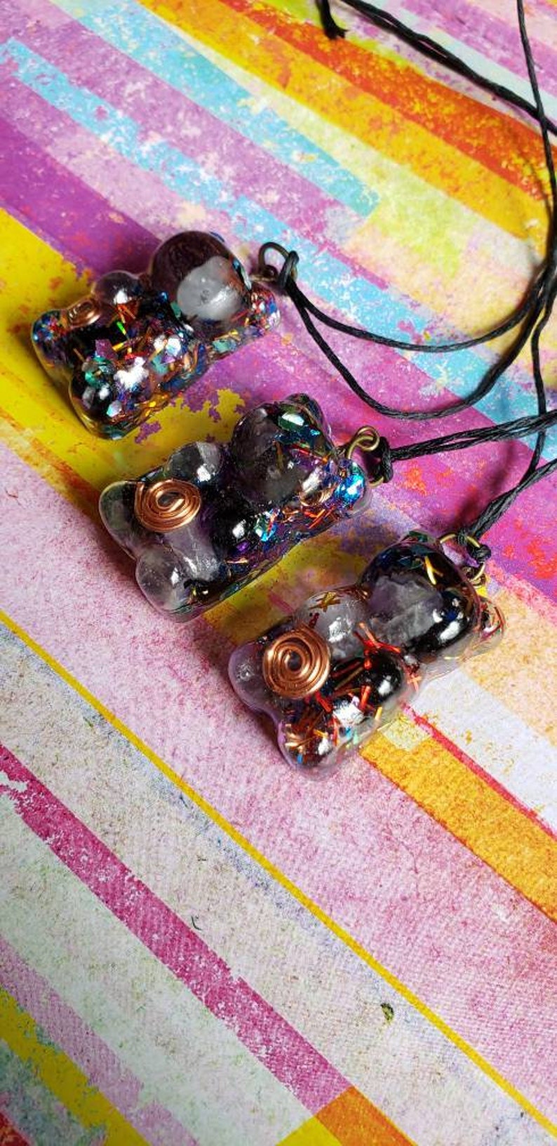 May include: Three resin bear pendants with black and clear crystal accents, copper wire, and iridescent glitter. The pendants are on a black cord and are displayed on a colorful striped background.