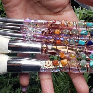 May include: Set of five makeup brushes with clear handles wrapped in copper wire and various colorful gemstones. The brushes have white bristles and silver metal ferrules.