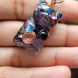 May include: A purple, black, and iridescent resin gummy bear charm with copper wire accents. The charm is attached to a black cord.