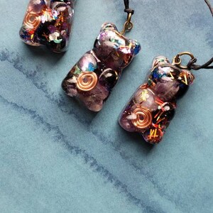 May include: Three purple resin bear pendants with gold and silver accents. Each pendant has a different arrangement of crystals and wire. The pendants are hanging from black cords.