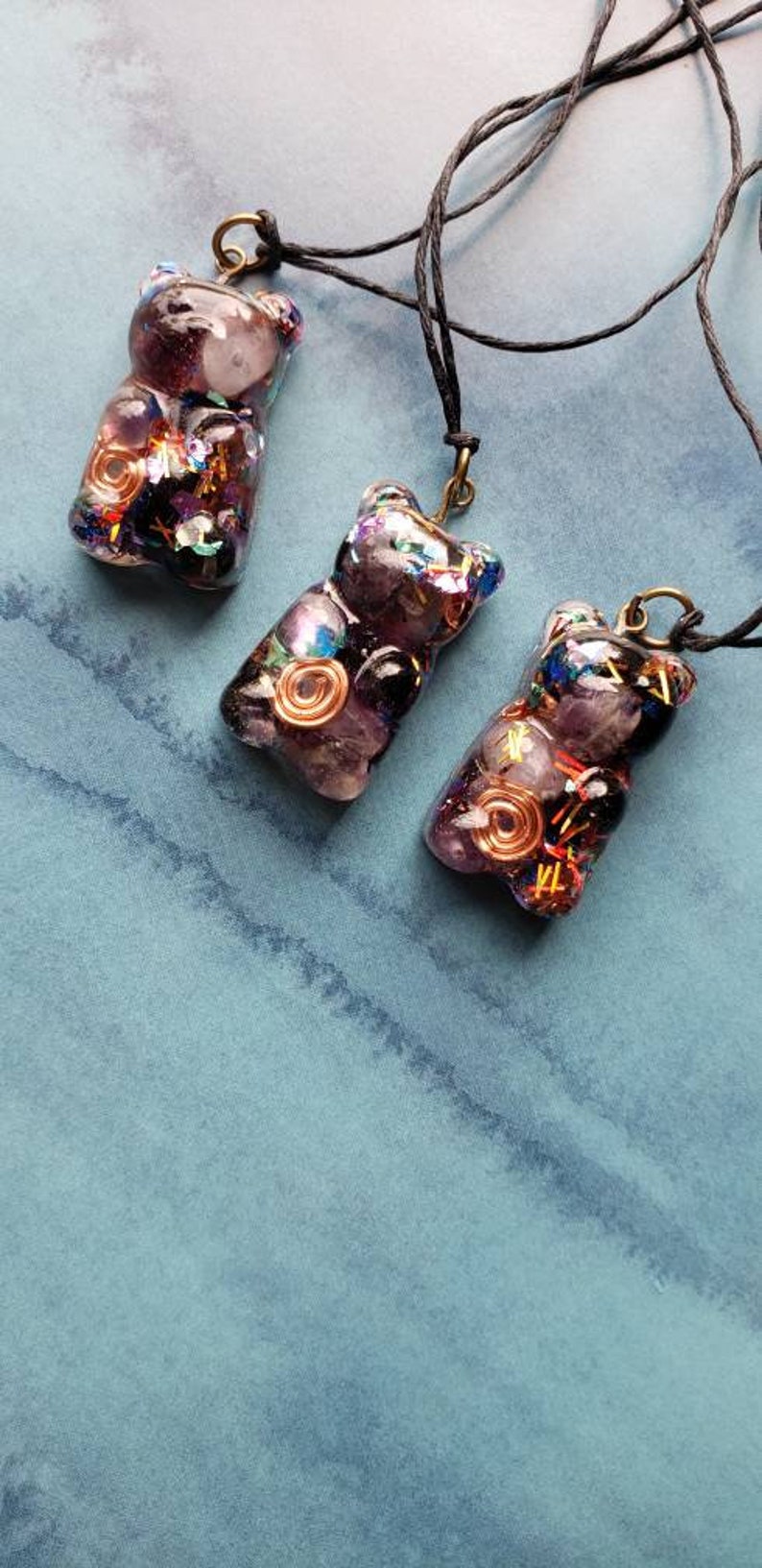 May include: Three purple resin bear pendants with gold and silver accents. Each pendant has a different arrangement of crystals and wire. The pendants are hanging from black cords.