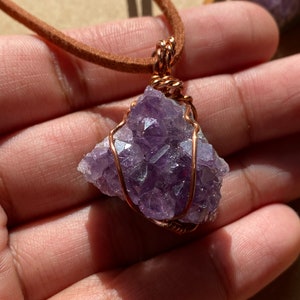 May include: A purple amethyst crystal wrapped in copper wire, hanging from a brown leather cord. The crystal is a cluster of small, pointed crystals.