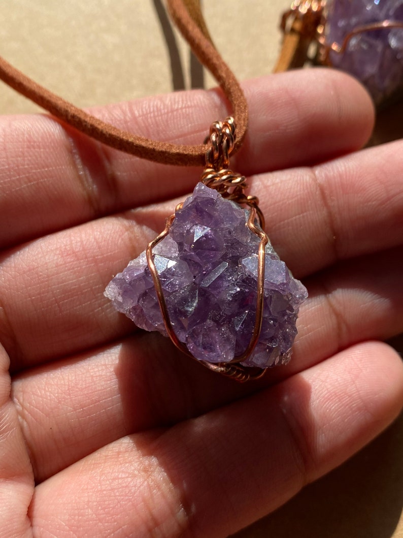 May include: A purple amethyst crystal wrapped in copper wire, hanging from a brown leather cord. The crystal is a cluster of small, pointed crystals.