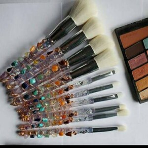 May include: A set of 10 makeup brushes with crystal and gemstone handles. The handles are wrapped in copper wire and feature a variety of colourful stones, including amethyst, turquoise, and citrine. The brushes have white bristles and are arranged in a fan shape.