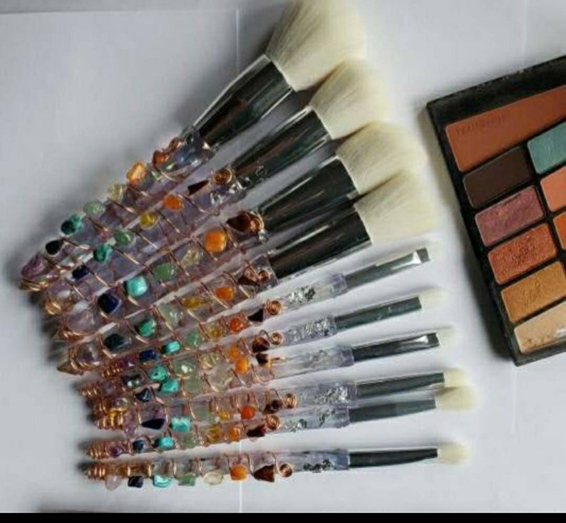 May include: A set of 10 makeup brushes with crystal and gemstone handles. The handles are wrapped in copper wire and feature a variety of colourful stones, including amethyst, turquoise, and citrine. The brushes have white bristles and are arranged in a fan shape.