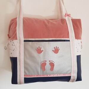 Peut inclure: Un sac à langer rose, bleu et blanc avec une poche zippée argentée et des motifs de mains et de pieds roses. Le sac a deux bretelles blanches et un nœud rose.