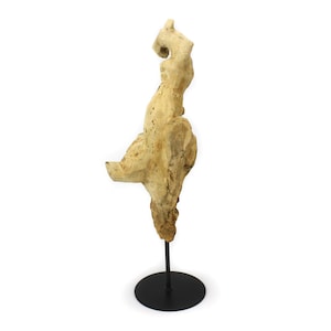 Pode incluir: Uma escultura abstrata de madeira clara, montada em um suporte de metal preto. A escultura tem uma forma natural e orgânica com uma textura áspera e um buraco perto do topo. O suporte tem uma base circular.