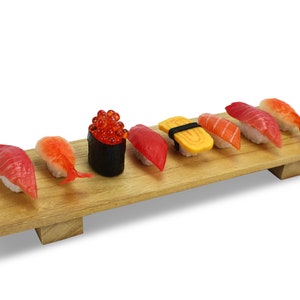 May include: A wooden serving board displays an assortment of sushi. The sushi includes nigiri with red tuna, salmon, shrimp, and a black seaweed roll topped with orange roe. The sushi is arranged on a light brown wooden board.