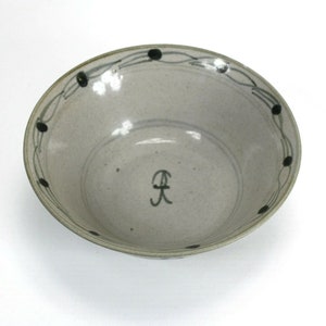Puede incluir: Un cuenco de cer&aacute;mica con un interior gris claro moteado. El borde presenta un patr&oacute;n decorativo de puntos negros y l&iacute;neas curvas. Una letra "R" negra estilizada est&aacute; pintada en el interior.