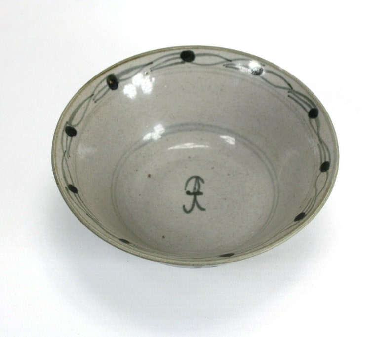 Puede incluir: Un cuenco de cer&aacute;mica con un interior gris claro moteado. El borde presenta un patr&oacute;n decorativo de puntos negros y l&iacute;neas curvas. Una letra "R" negra estilizada est&aacute; pintada en el interior.