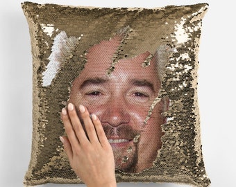 Può includere: Un cuscino di paillettes dorate con un viso d'uomo che spunta da un buco nelle paillettes. L'uomo sorride e ha la barba.