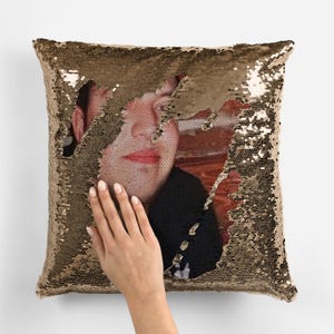 May include: A gold sequin pillow with a photo of a person's face peeking through the sequins.