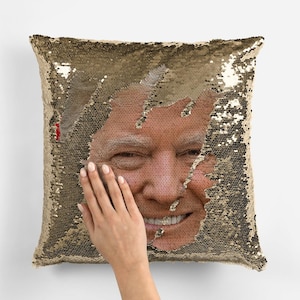 May include: A gold sequin pillow with a smiling face peeking out from the sequins. The face is a close-up of a man with brown hair and a light complexion.