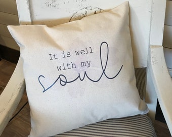 May include: A white pillow with black text that reads "It is well with my soul". The pillow is sitting on a white chair with a striped cushion.