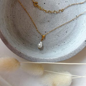 Puede incluir: Un delicado collar dorado con un colgante de perla única en forma de lágrima. El collar se exhibe en un cuenco gris texturizado, con elementos decorativos beige suaves y esponjosos en primer plano. La cadena y el cierre son visibles.