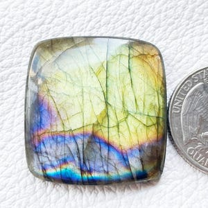 Puede incluir: Gema de labradorita de corte cuadrado con colores iridiscentes de azul, verde y amarillo. La piedra tiene un aspecto agrietado y texturizado. Una moneda de veinticinco centavos estadounidense está al lado para comparar el tamaño.