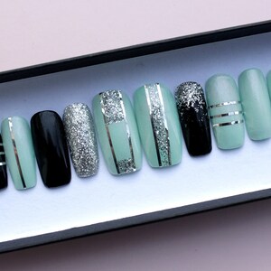 Puede incluir: Un conjunto de diez u&ntilde;as postizas con una base verde menta y detalles de purpurina plateada. Algunas u&ntilde;as tienen rayas plateadas, mientras que otras tienen una base negra con purpurina plateada.