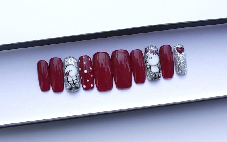 May include: Set of ten red press-on nails with various designs. Some nails have white polka dots, others have a silver glitter finish, and one nail features a cartoon couple with a heart.