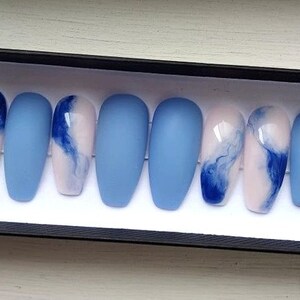 Puede incluir: Un juego de diez u&ntilde;as artificiales en una caja negra. Las u&ntilde;as alternan entre azul claro mate liso y un dise&ntilde;o de blanco y rosa con remolinos azules. Las u&ntilde;as tienen forma ovalada.