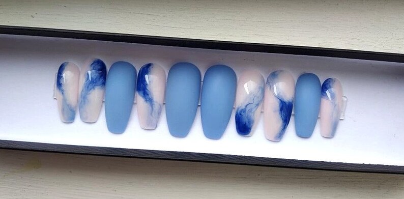 Puede incluir: Un juego de diez u&ntilde;as artificiales en una caja negra. Las u&ntilde;as alternan entre azul claro mate liso y un dise&ntilde;o de blanco y rosa con remolinos azules. Las u&ntilde;as tienen forma ovalada.