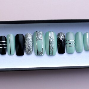 Puede incluir: Un conjunto de diez u&ntilde;as postizas con un dise&ntilde;o de purpurina verde menta, negro y plateado. Las u&ntilde;as est&aacute;n dispuestas en una caja.