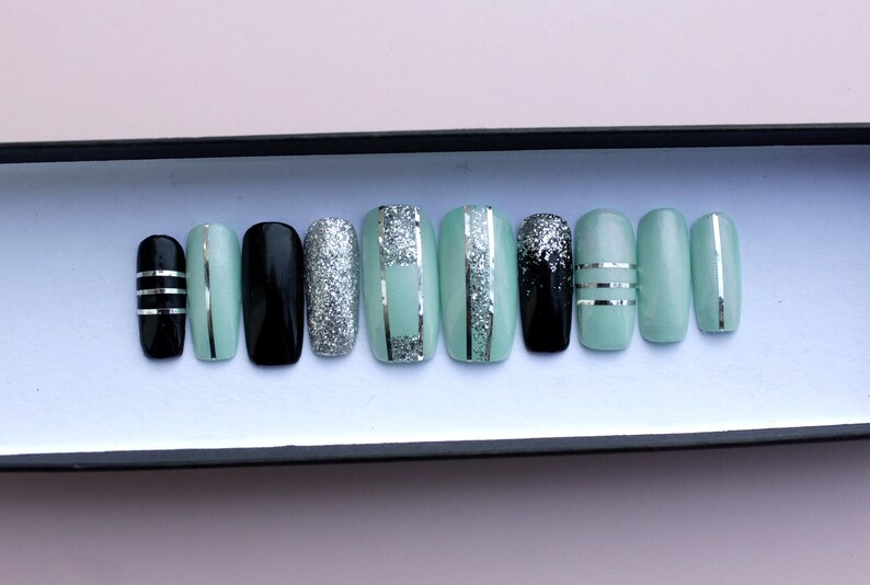 Puede incluir: Un conjunto de diez u&ntilde;as postizas con un dise&ntilde;o de purpurina verde menta, negro y plateado. Las u&ntilde;as est&aacute;n dispuestas en una caja.