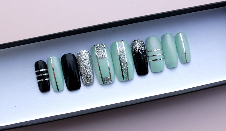 Puede incluir: Un conjunto de diez u&ntilde;as postizas con una base verde menta y detalles de purpurina plateada. Algunas u&ntilde;as tienen rayas plateadas, mientras que otras tienen una base negra con purpurina plateada.