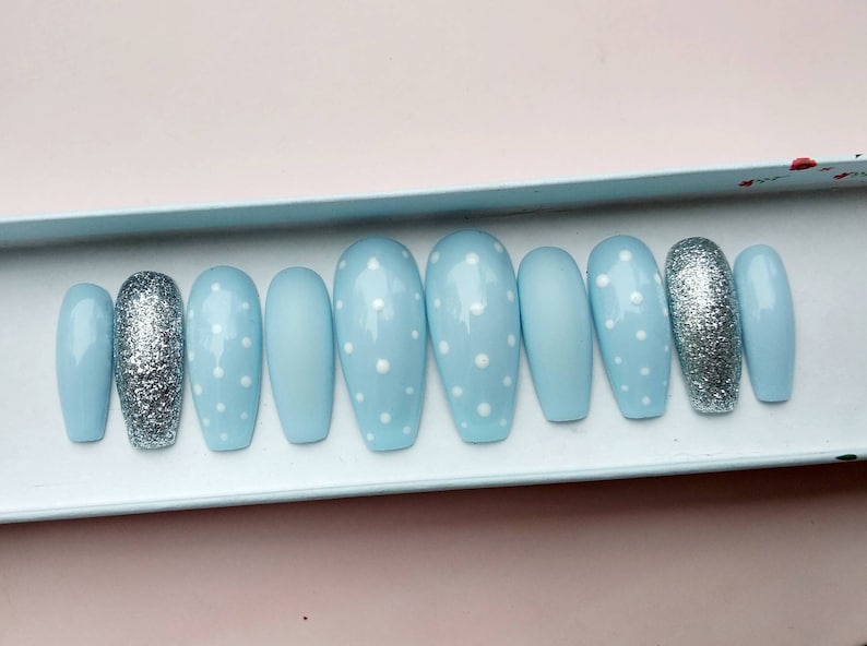 K&ouml;nnte beinhalten: Ein Set mit zehn hellblauen, sargf&ouml;rmigen Press-on-N&auml;geln mit wei&szlig;en Punkten. Ein Nagel ist silberfarben glitzernd.