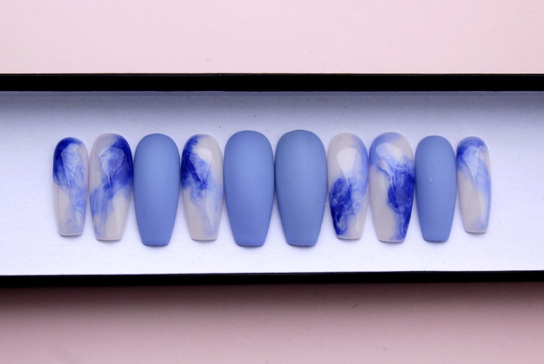 Puede incluir: Un juego de diez u&ntilde;as artificiales en forma de ata&uacute;d. Las u&ntilde;as alternan entre azul mate s&oacute;lido y un dise&ntilde;o de remolinos azules sobre una base blanca. Las u&ntilde;as se exhiben en una caja negra.