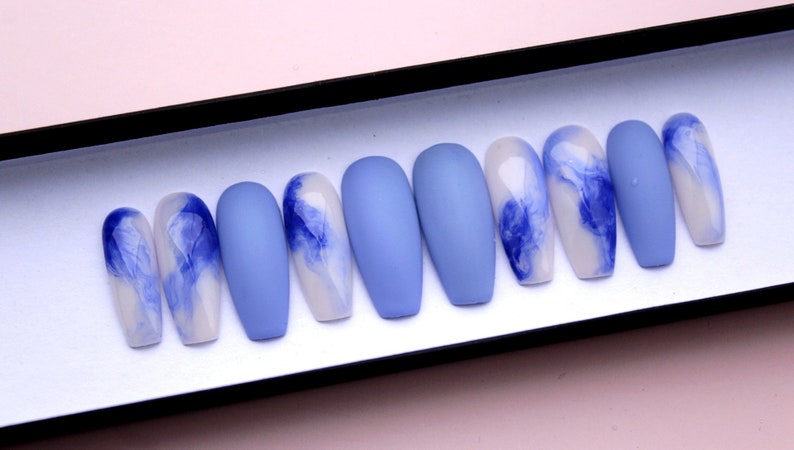 Puede incluir: Un juego de diez u&ntilde;as artificiales en una caja expositora. Las u&ntilde;as alternan entre azul claro mate liso y blanco con dise&ntilde;os veteados azules. Las u&ntilde;as son de forma ovalada y var&iacute;an en tama&ntilde;o.
