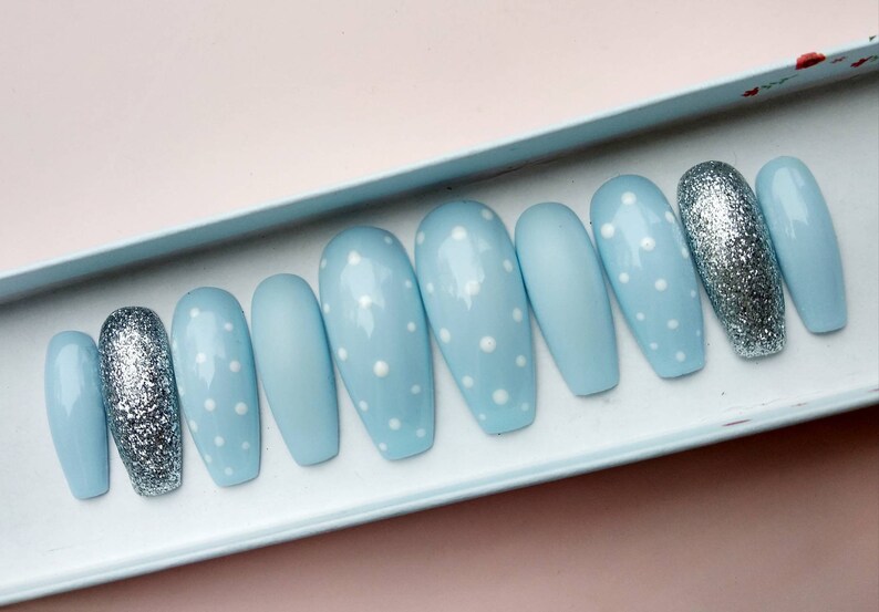 K&ouml;nnte beinhalten: Ein Set mit zehn hellblauen, sargf&ouml;rmigen Press-on-N&auml;geln mit wei&szlig;en Punkten. Zwei der N&auml;gel haben silbernen Glitzer.