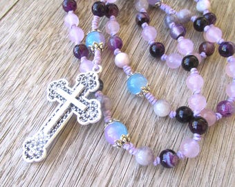 May include: A purple, pink, and blue beaded rosary with a silver cross pendant. The rosary is made with natural stones and is strung on a purple cord.