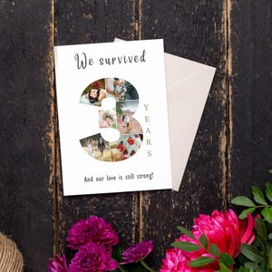 Pode incluir: Um cart&atilde;o de anivers&aacute;rio branco com as palavras "We survived" e "And our love is still strong!" impressas. O n&uacute;mero "3" &eacute; preenchido com fotos. Um envelope bege e flores cor-de-rosa s&atilde;o vis&iacute;veis.