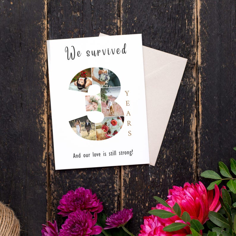 Pode incluir: Um cart&atilde;o de anivers&aacute;rio branco com as palavras "We survived" e "And our love is still strong!" impressas. O n&uacute;mero "3" &eacute; preenchido com fotos. Um envelope bege e flores cor-de-rosa s&atilde;o vis&iacute;veis.