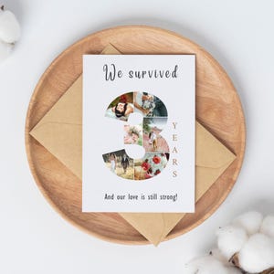 Pode incluir: Um cart&atilde;o branco com o texto "We survived" e "3 YEARS" num design estilizado. O n&uacute;mero 3 &eacute; preenchido com v&aacute;rias fotos pequenas. O cart&atilde;o est&aacute; numa bandeja de madeira com um envelope castanho.