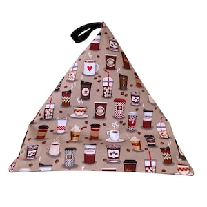 May include: A triangular beige cushion with a black handle. The cushion is covered in a fabric with a pattern of coffee cups and coffee beans.