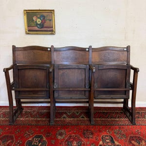 May include: A three-seat wooden theatre bench featuring dark brown wood, folding seats, and carved floral detailing on the upper backrests. The bench has curved armrests on the ends and a sturdy frame, positioned above a red patterned rug.