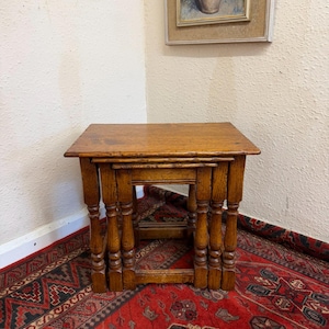 May include: A set of three nested tables crafted from medium-brown wood. Each table features a rectangular top with rounded edges and four turned legs connected by horizontal stretchers near the base, allowing them to slide neatly together.