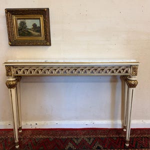 May include: A rectangular console table with a white marble top, cream-coloured frame, and gold-painted carved details on the legs and apron. Above the table hangs a small landscape painting in a thick, ornate, dark gold-toned wooden frame.