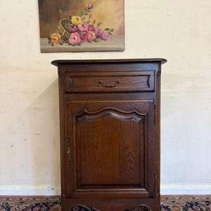 May include: A dark oak cabinet with a drawer and a door, featuring decorative carvings and brass handles. Above, a painting depicts a basket of pink, yellow, and orange roses. The cabinet is a classic piece of furniture.