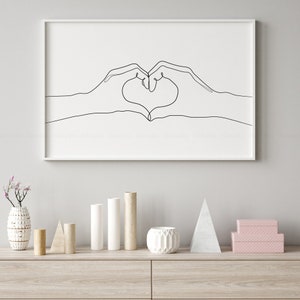 May include: A framed minimalist art print featuring a continuous line drawing of two hands forming a heart shape. The artwork is in black against a white background, encased in a white frame. The print is displayed on a light-colored wooden surface.