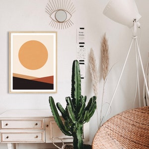 May include: A framed art print featuring a minimalist landscape design. The artwork showcases a large orange sun against a cream-colored sky, with layered brown and black shapes representing hills. The print is in a light wood frame.