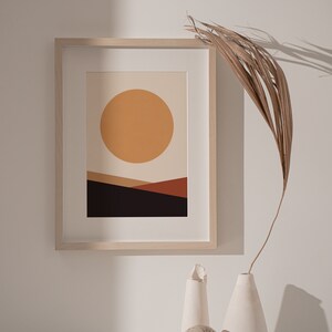 May include: A framed abstract art print featuring a large orange sun over a landscape of brown and black geometric shapes. The artwork is in a light wood frame and is displayed with white vases and a dried palm frond.