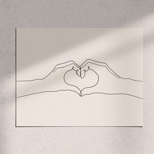 May include: A minimalist line art print featuring two hands forming a heart shape. The artwork is rendered in a continuous black line on a cream-colored background. The print is displayed on a wall with natural light and shadows.