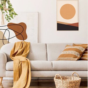 May include: A living room scene featuring a light gray sofa with a mustard yellow waffle-weave throw blanket. Decorative pillows with geometric patterns and a woven basket add texture. Artwork with abstract designs and a framed landscape print complete the decor.