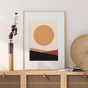 May include: Framed abstract art print featuring a large orange sun over a landscape of brown and black hills. The artwork is in a gold frame with a white mat. The print is displayed on a wooden cabinet with a vase and decorative items.