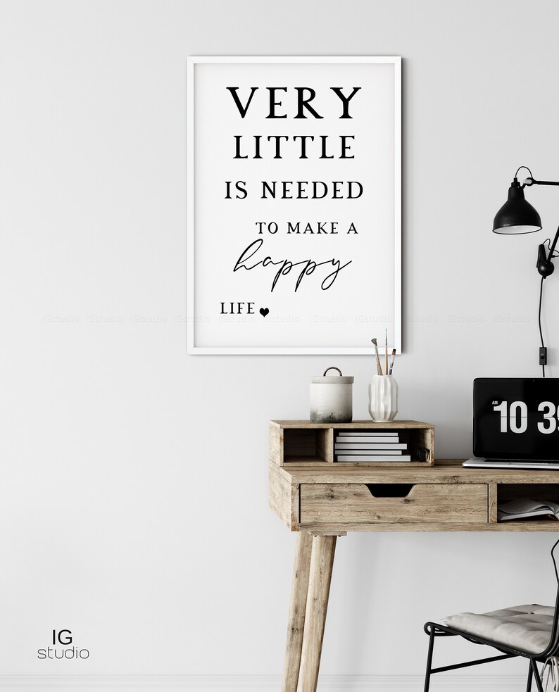 May include: A framed print with the text "VERY LITTLE IS NEEDED TO MAKE A happy LIFE" in black font. The print hangs above a wooden desk with a laptop, lamp, and decorative items.