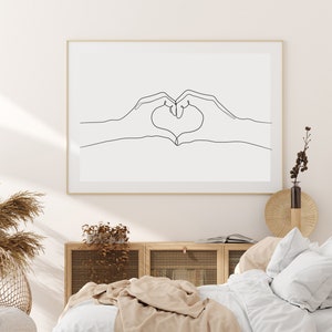 May include: A framed art print featuring a minimalist line drawing of two hands forming a heart shape. The artwork is in black against a white background, set within a gold frame. The print is displayed in a bedroom setting with neutral tones.