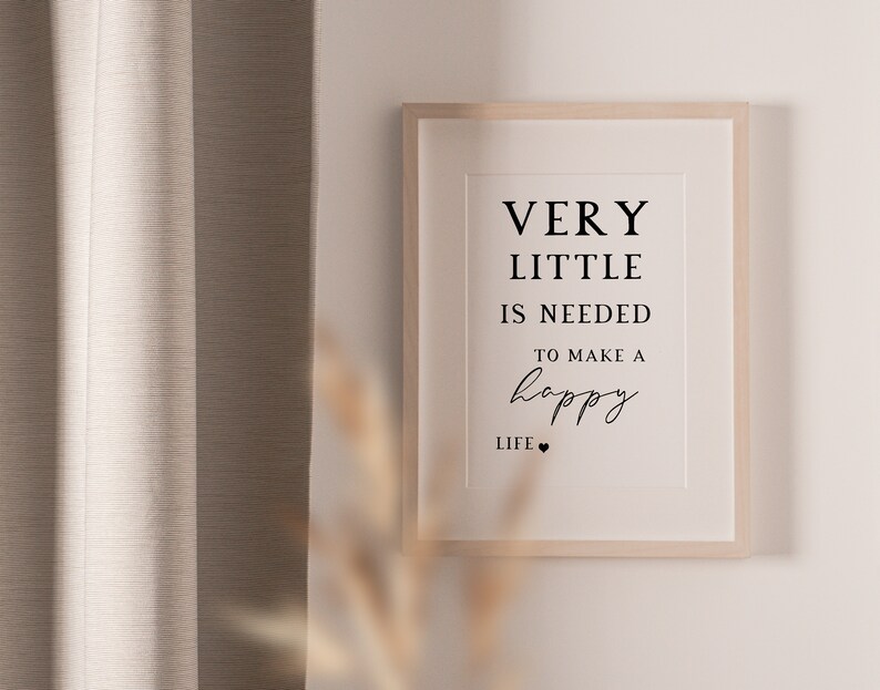 May include: A framed print with the text "VERY LITTLE IS NEEDED TO MAKE A happy LIFE" in black font. The frame is light wood, and the background is a neutral color. The print is hanging on a white wall.