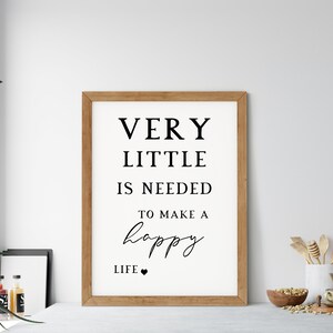 May include: A framed print with the text "VERY LITTLE IS NEEDED TO MAKE A happy LIFE" in black on a white background. The frame is made of light brown wood. The print is displayed on a table with kitchen utensils and decor.